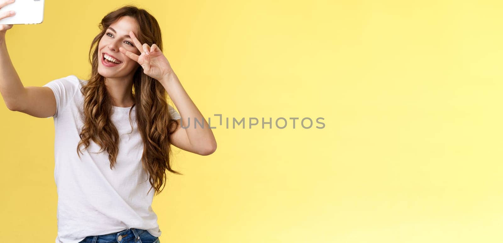 Joyful charismatic good-looking curly-haired woman white t-shirt show peace victory sign smiling broadly posing tender friendly expression smartphone front camera take selfie yellow background. Lifestyle.