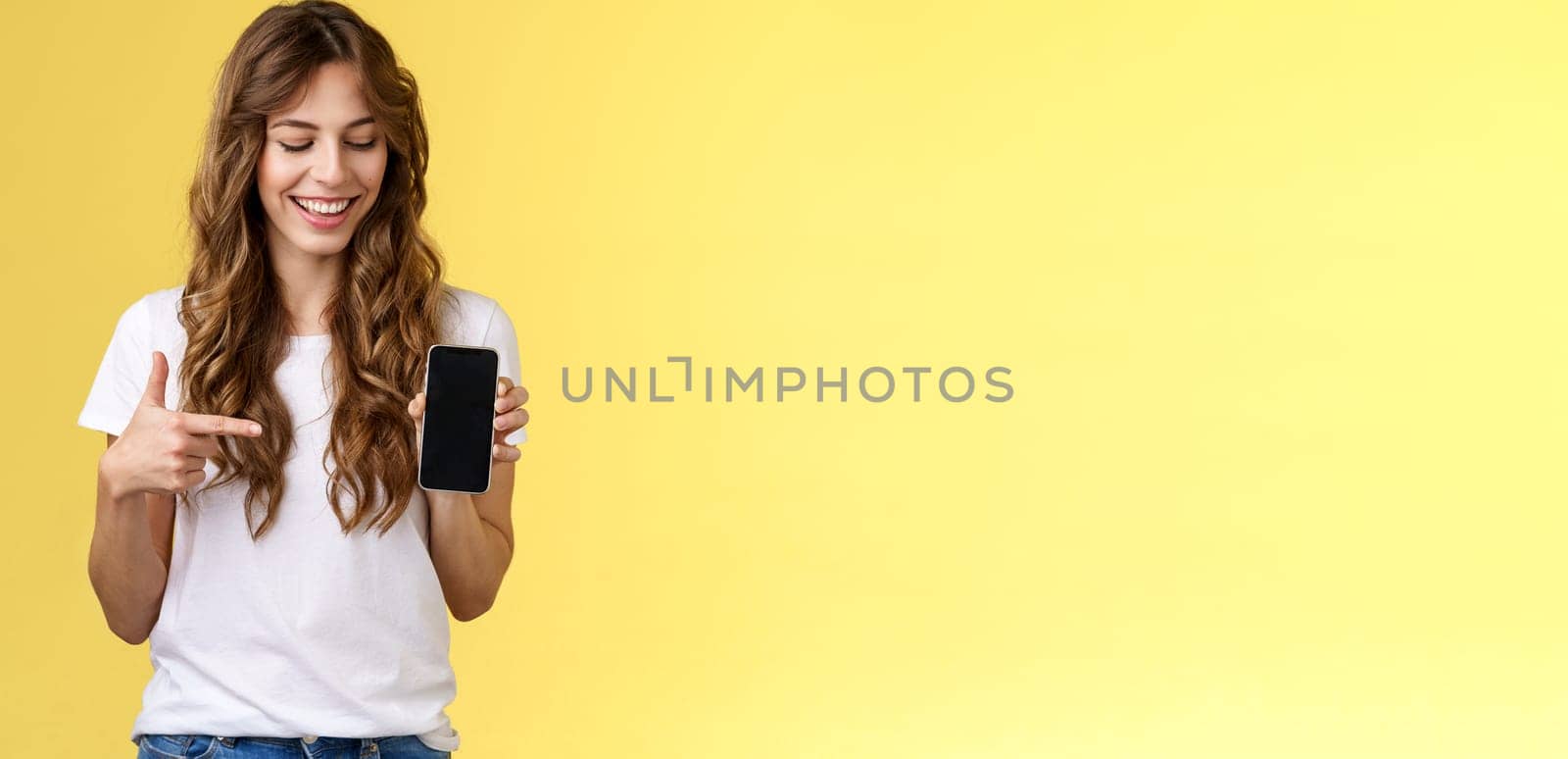 Intrigued charismatic happy young female blogger show what photo app use edit social media profile hold smartphone looking pointing index finger mobile phone blank screen smiling amused by Benzoix