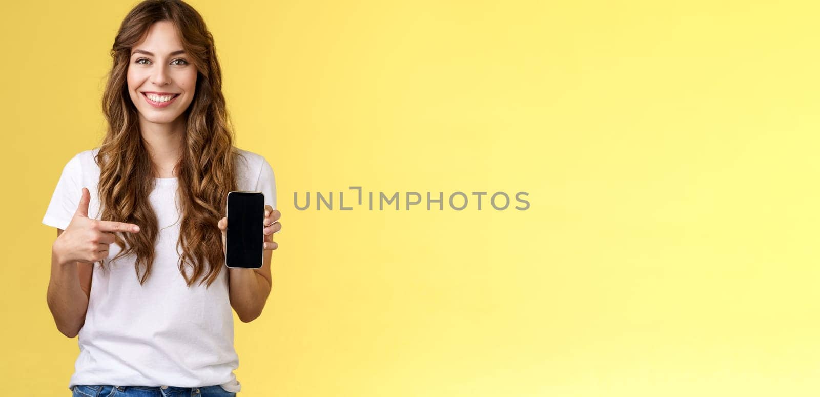 Girl have everything need smartphone. Cheerful relaxed carefree woman long curly hairstyle hold mobile phone pointing forefinger cellphone smiling broadly explain how app works yellow background.