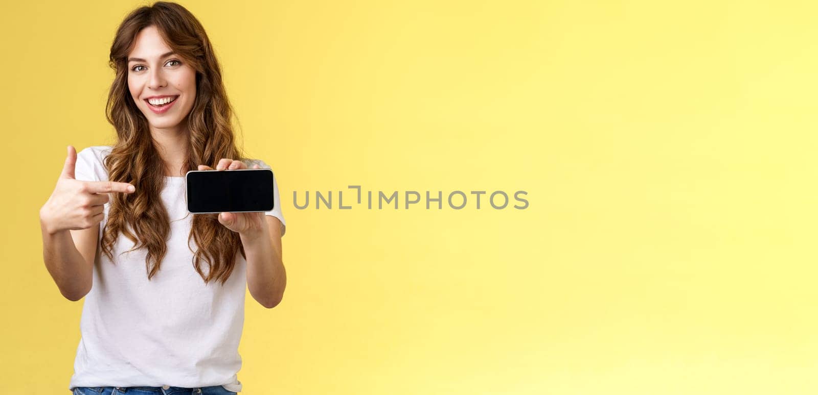 Confident sassy good-looking stylish woman introduce awesome game app show smartphone pointing index finger blank mobile phone screen smiling delighted recommend try application yellow background by Benzoix