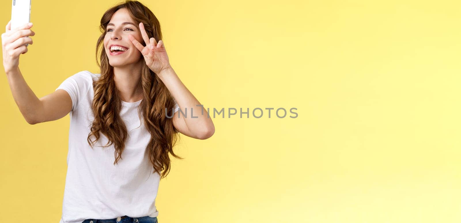 Positive charisamtic happy young girl curly-haired raise smartphone taking selfie show peace victory sign having video call stand yellow background smiling mobile front camera posing lively. Lifestyle.