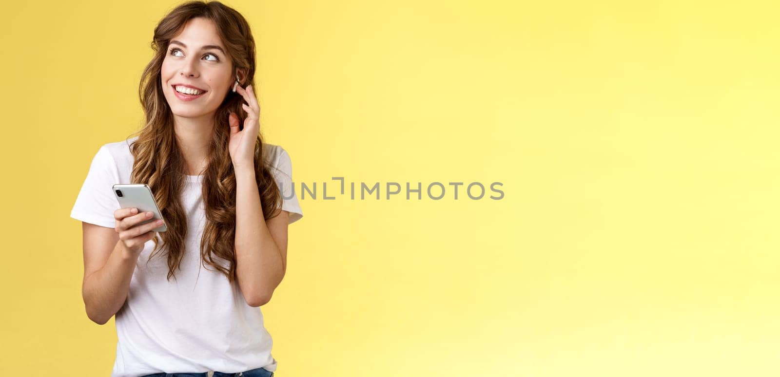 Dreamy happy cheerful curly-haired girl look around contemplate beautiful summer weather listening music touch wireless earbud calling friend talking via earphones hold smartphone yellow background. Lifestyle.