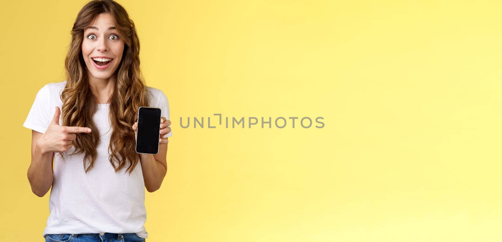 Surprised happy lucky girl winning online internet lottery smiling broadly hold smartphone pointing blank mobile phone screen showing display grinning excited enthusiastic stand yellow background.