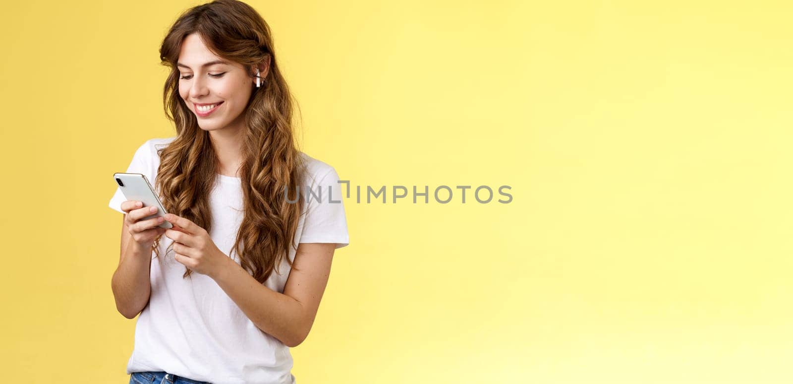 Happy caring tender feminine girlfriend curly long hair hold smartphone picking song listen way home smiling broadly look mobile phone scroll music platform wear wireless earbud yellow background by Benzoix
