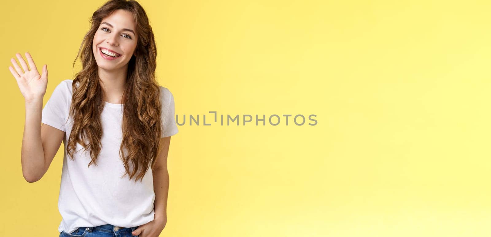 Modest cute friendly outgoing attractive european woman long curly haircut waving palm sociable conversation introduce herself smiling broadly say hi hello greeting coworker yellow background.