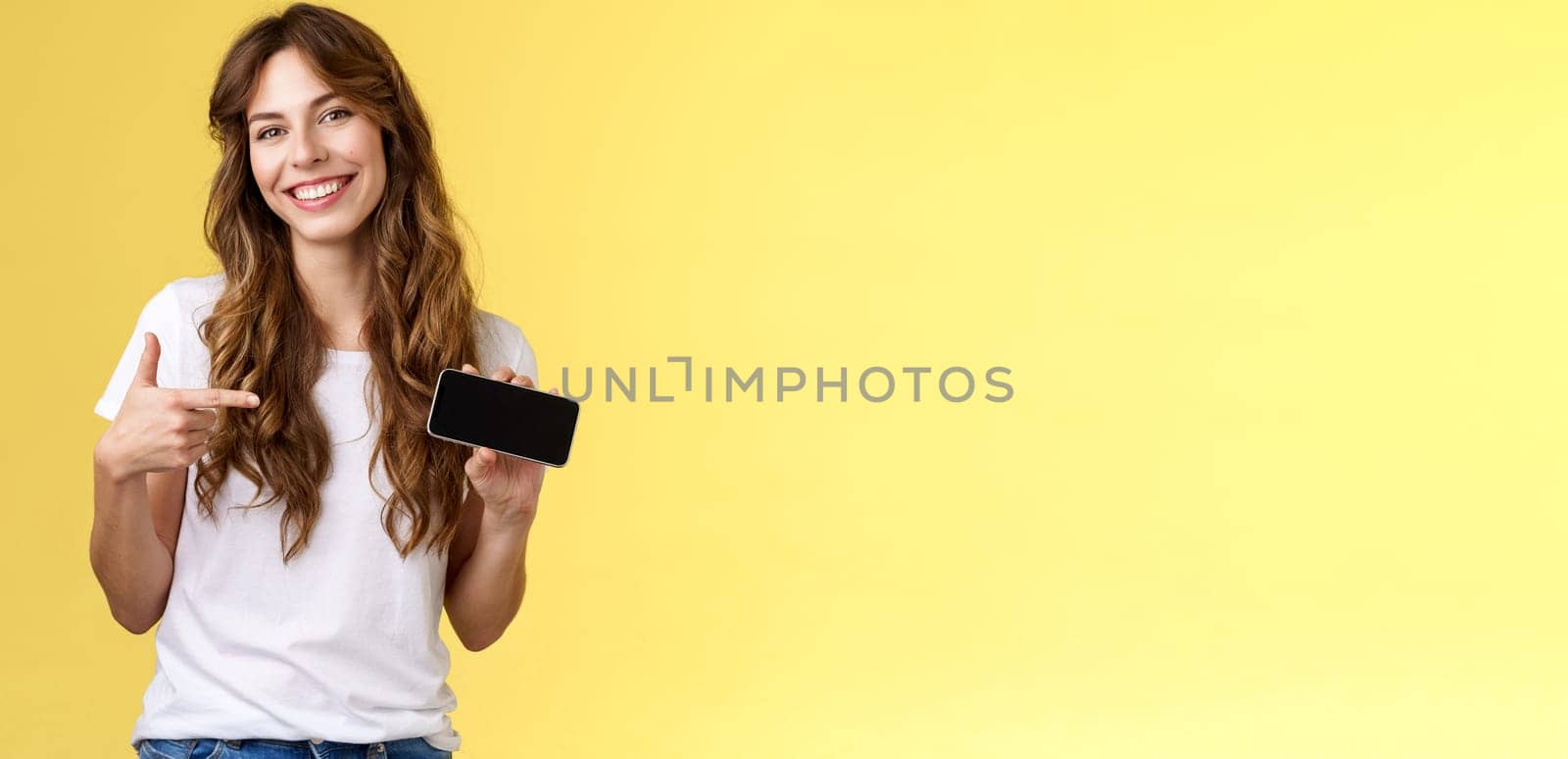 Cheerful good-looking friendly carefree young girl hold smartphone horizontal pointing index finger mobile phone screen smiling broadly introduce awesome game app stand yellow background.