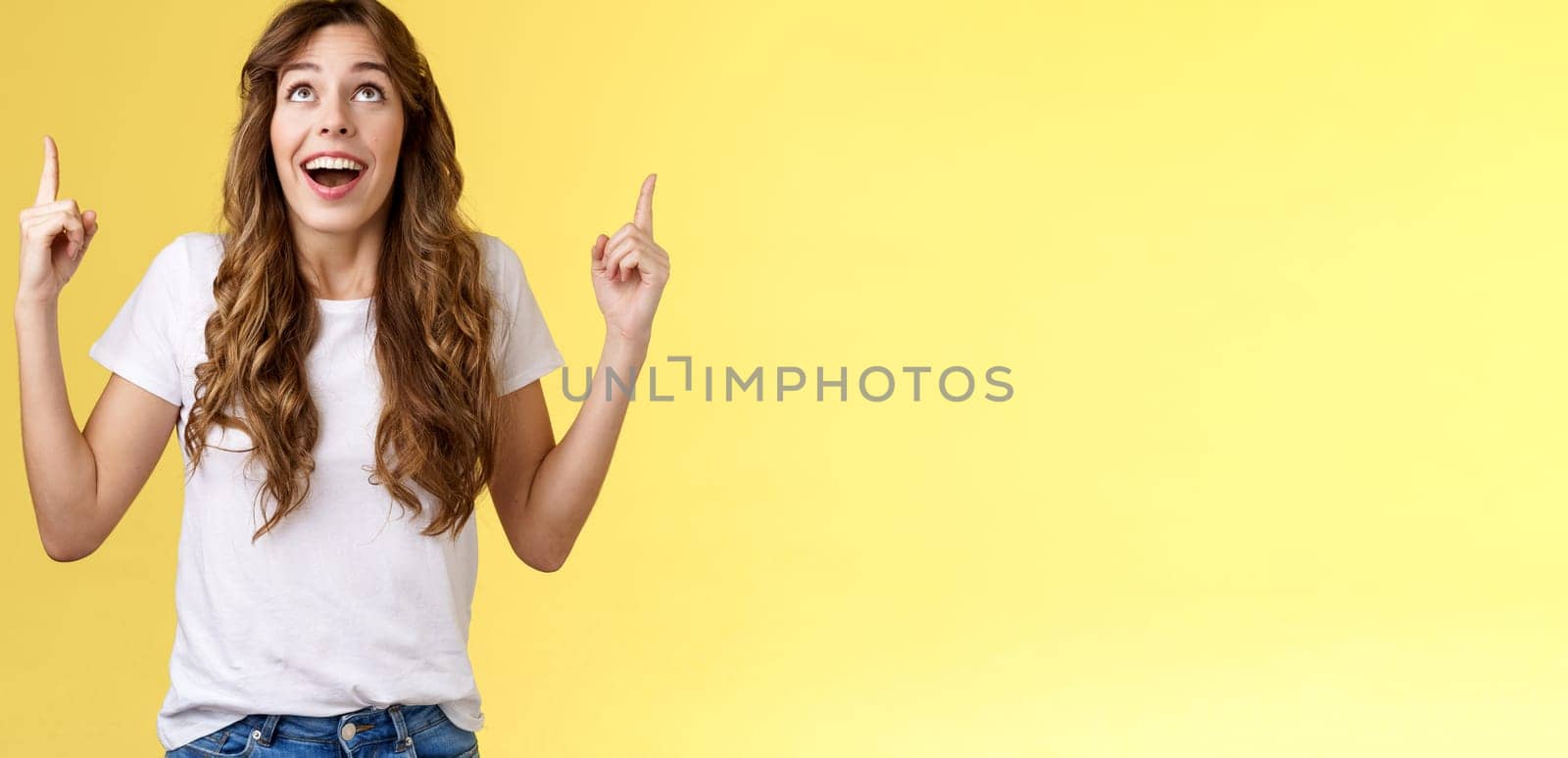 Happy impressed attractive girl long curly haircut drop jaw amused smiling surprised delighted look pointing up index fingers stare admiration fascinated awesome promo offer yellow background.Lifestyle.