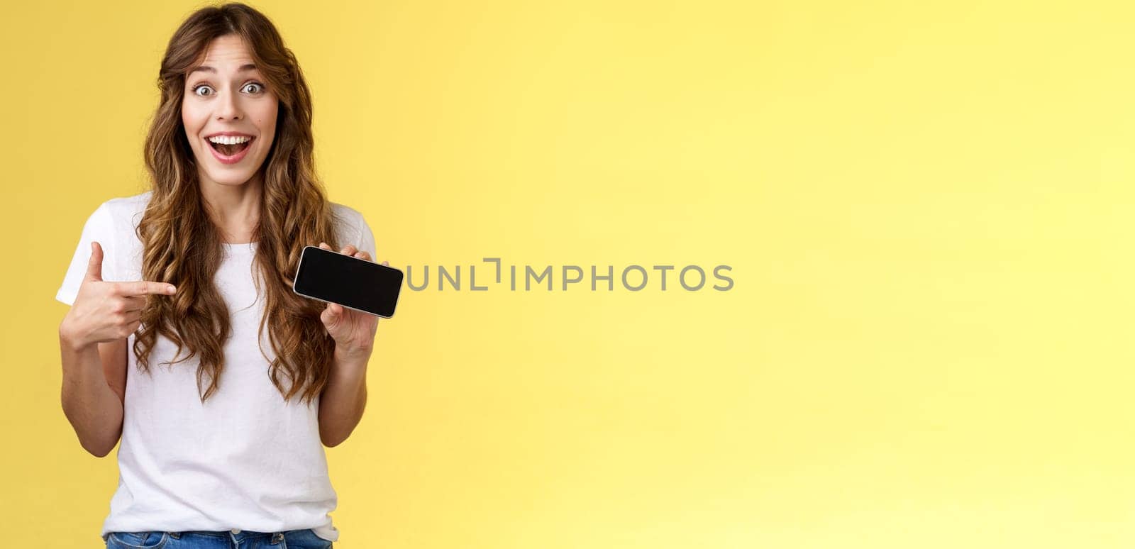 Cheerful surprised cute lucky girl beat best score awesome game showing smartphone display pointing index finger horizontal screen mobile phone introduce cool app smiling broadly yellow background. Lifestyle.