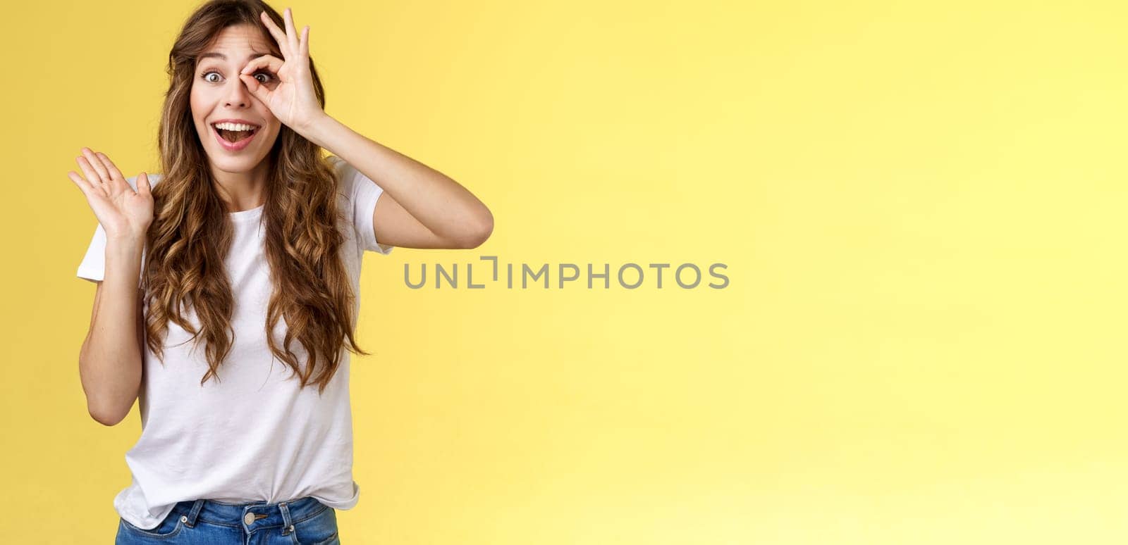 Oh hi I see you. Fascinated cute charming young girl waving palm say hello impressed look through okay ring gesture impressed finally fixed sight stand astonished amazed yellow background.