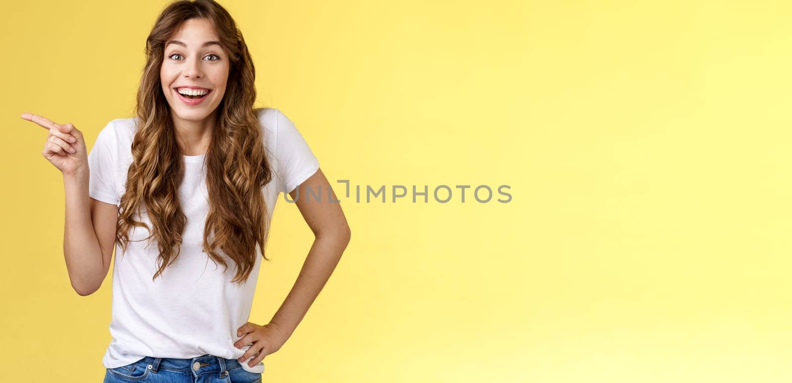 Lively surprised happy enthusiastic good-looking woman long curly hairstyle white t-shirt laughing impressed astonished pointing left index finger discuss interesting exhibition yellow background.