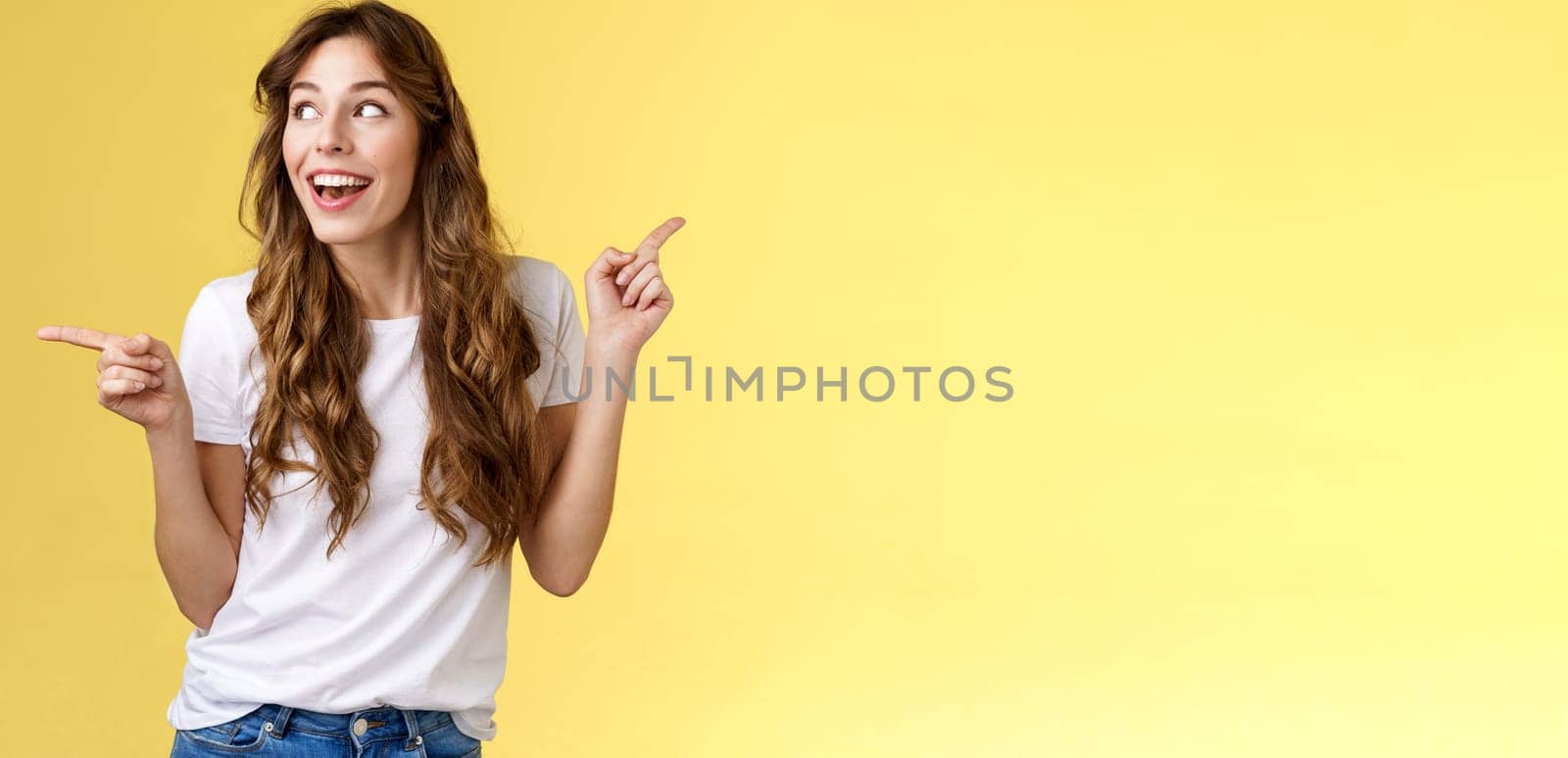 Amused cheerful smiling happy young curly-haired woman chestnut hair observe curiously copy space grinning admiration joy pointing sideways making choice impressed satisfied turn left.
