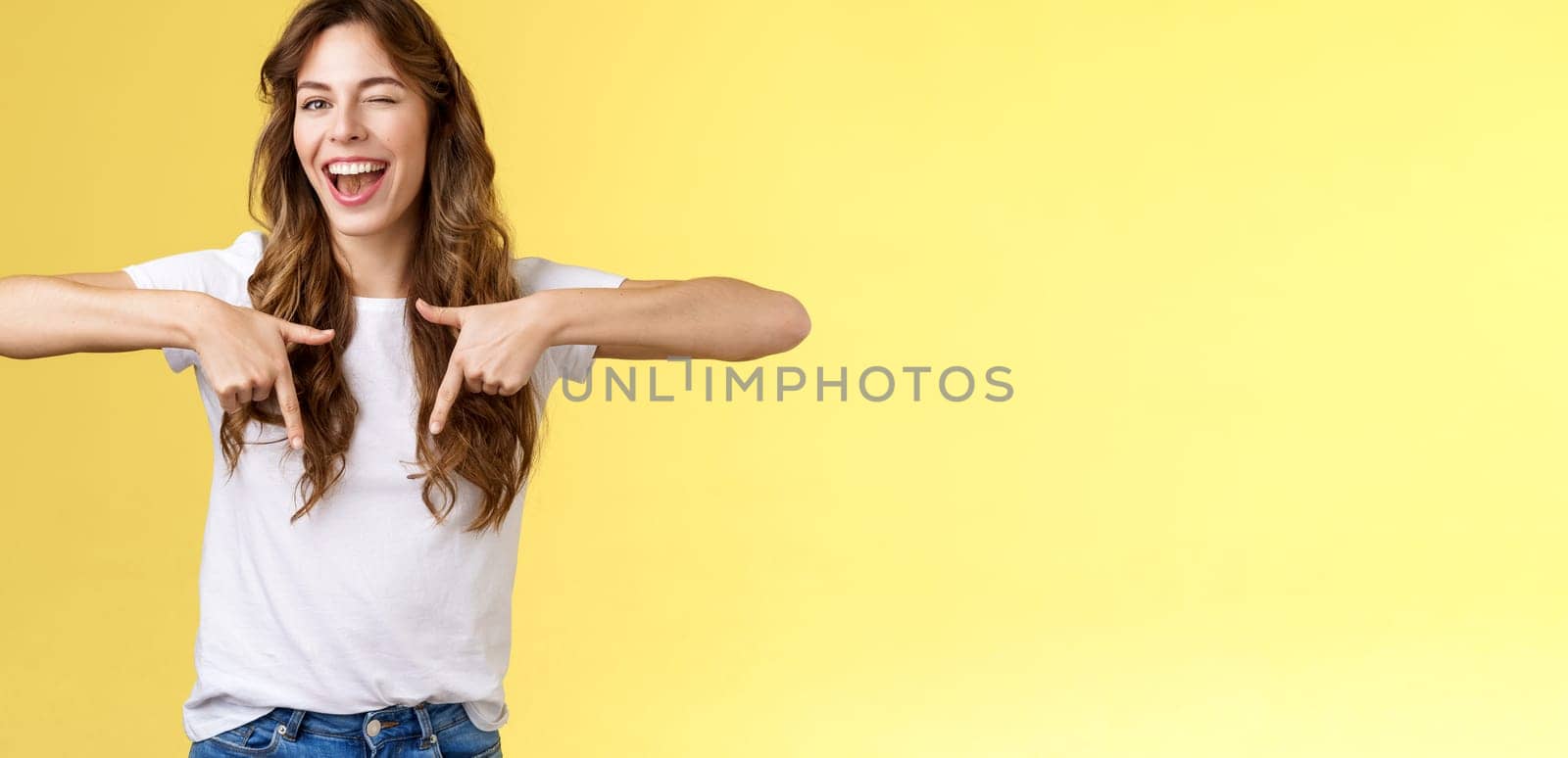 Hey visit my link would not regret. Lively cheeky enthusiastic woman curly long haircut wink sassy smiling broadly show you perfect copy space pointing down bottom promo yellow background.