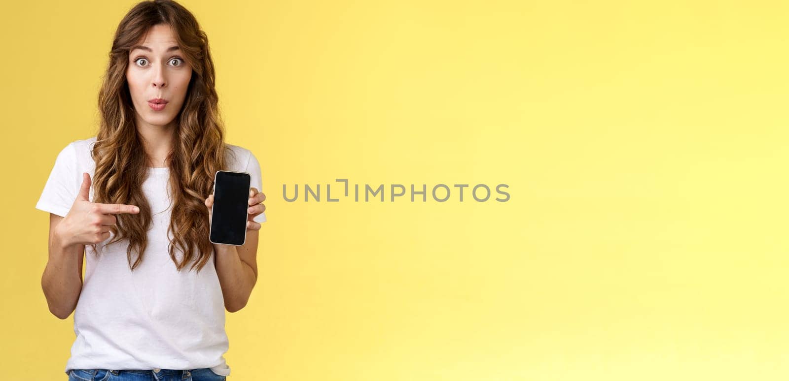 Intriguing app check out. Enthusiastic surprised attractive girlfriend gossiping friend new boyfriend showing curious photo smartphone hold mobile phone pointing telephone screen yellow background.