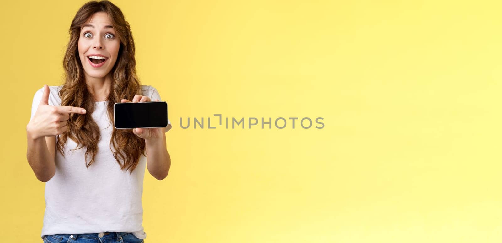 Impressed upbeat happy lucky girl curly long hairstyle open mouth admiration joy like awesome new app show smartphone screen horizontal phone display stand yellow background amazed by Benzoix