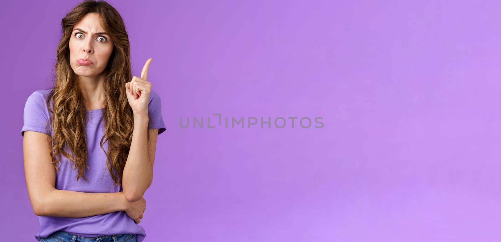 Strict funny curly-haired girl pulling disappointed angry face popping eyes stare camera shaking index finger disapproval scold bad behaviour stand serious bossy purple background.