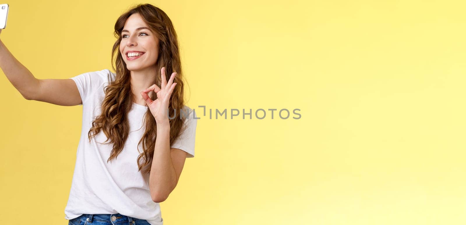 Stylish carefree urban confident curly-haired girl recording video blog followers extend arm hold smartphone taking selfie front camera show okay ok approval gesture yellow background by Benzoix