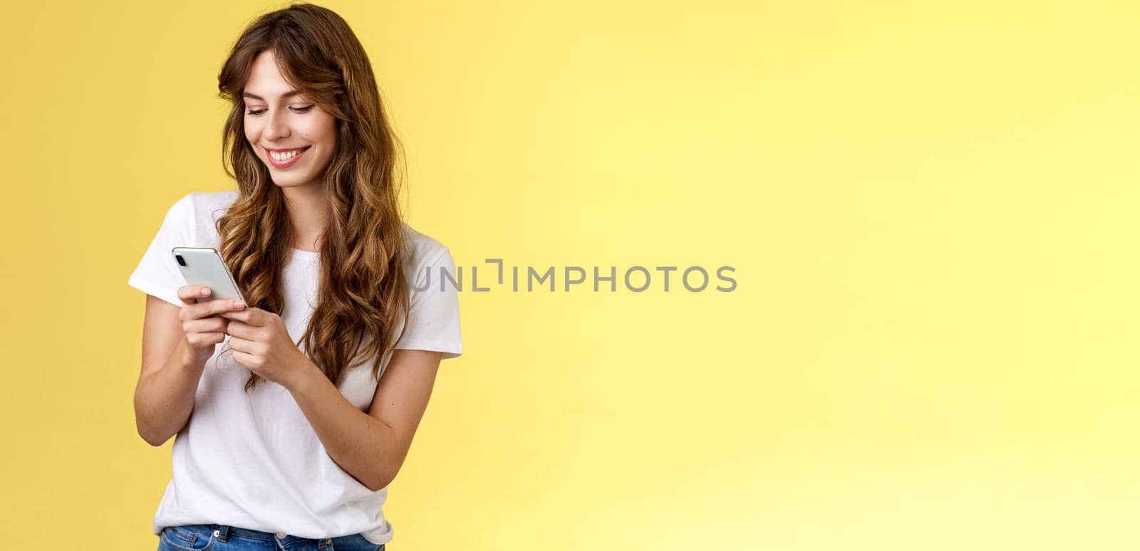 Cheerful lovely girlfriend texting friend pleased cute smile tap smartphone screen smiling broadly look mobile phone display tenderly writing post contemplate touching photo yellow background.