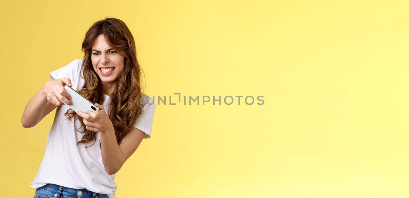 Girl must win race. Excited determined focused eager playful woman hold horizontally smartphone tilt body playing intense game frowning grimacing stare mobile phone screen stand yellow background.