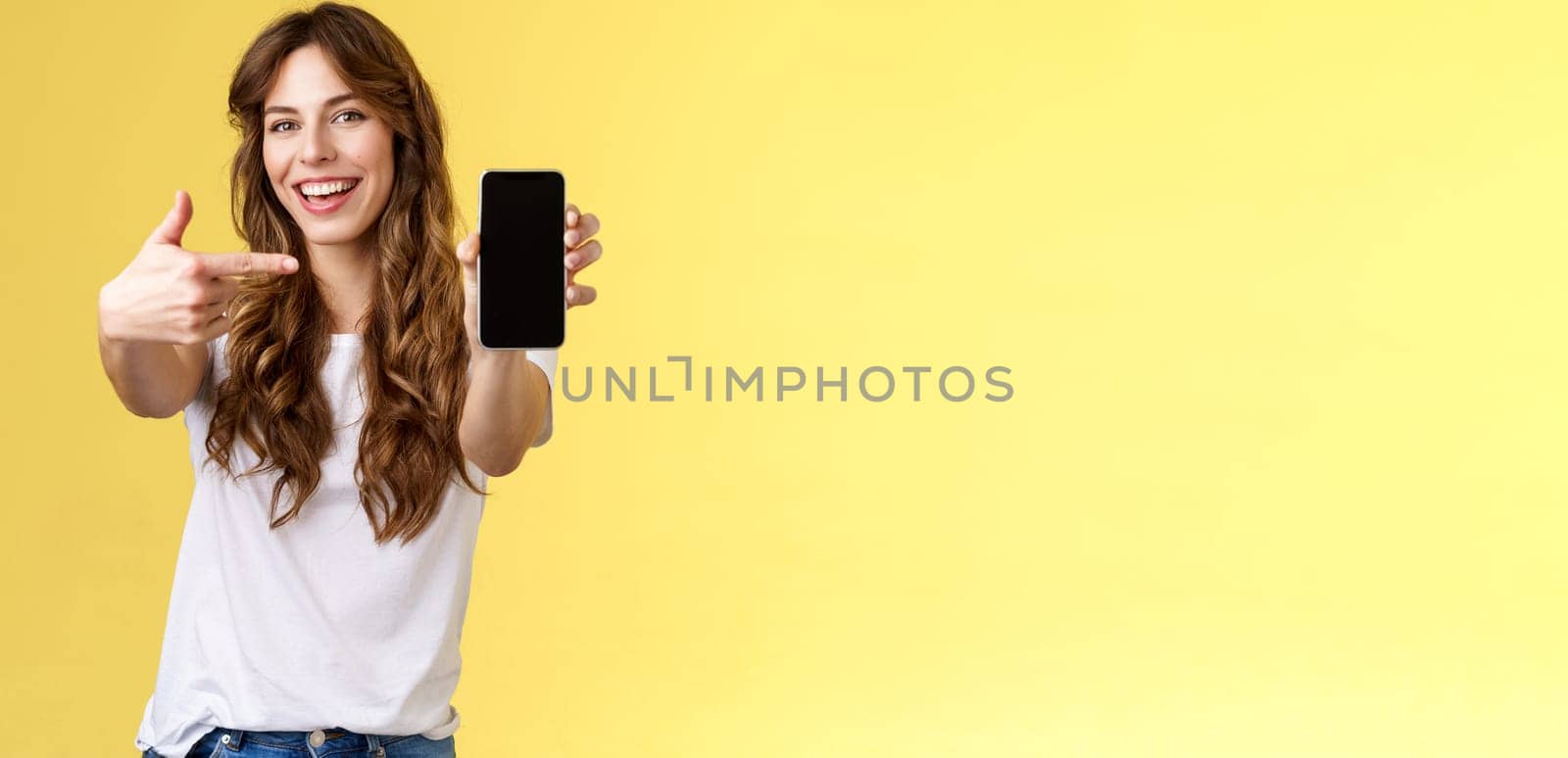 Cheerful sassy good-looking sociable girl curly long hair extend arm holding smartphone pointing index finger mobile phone screen smiling broadly recommend cool app blogger promote social page. Lifestyle.