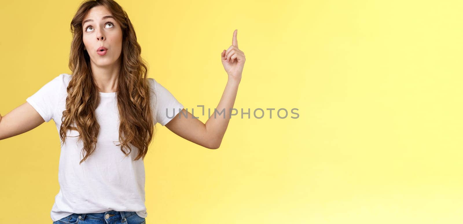 Impressed curious wondered attractive european woman long curly haircut look pointing index fingers up top copy space promo react astonished fascinated surprising event yellow background.