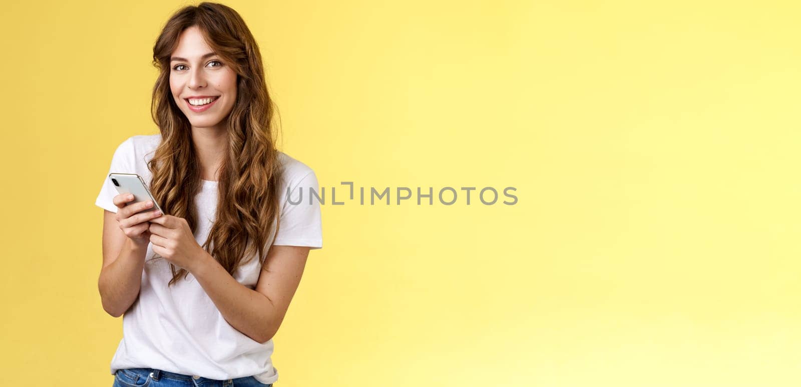 Enthusiastic good-looking urban girl wear white t-shirt standing casual smiling delighted camera texting hold smartphone scroll social media feed stand yellow background browsing network. Lifestyle.