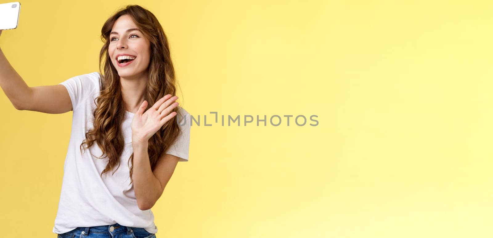 Friendly lively good-looking cheerful feminine girl extend arm hold smartphone record video blog waving front camera smiling broadly talking fans videocalling taking selfie yellow background. Lifestyle.