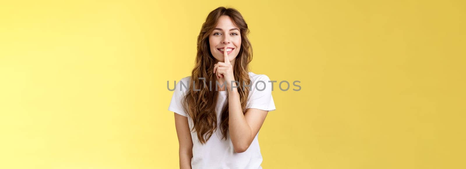 Cute cunning lovely european girl curly hairstyle hiding beauty secret smiling sensually show hush shush gesture index finger pressed lips grinning joyfully stand yellow background.