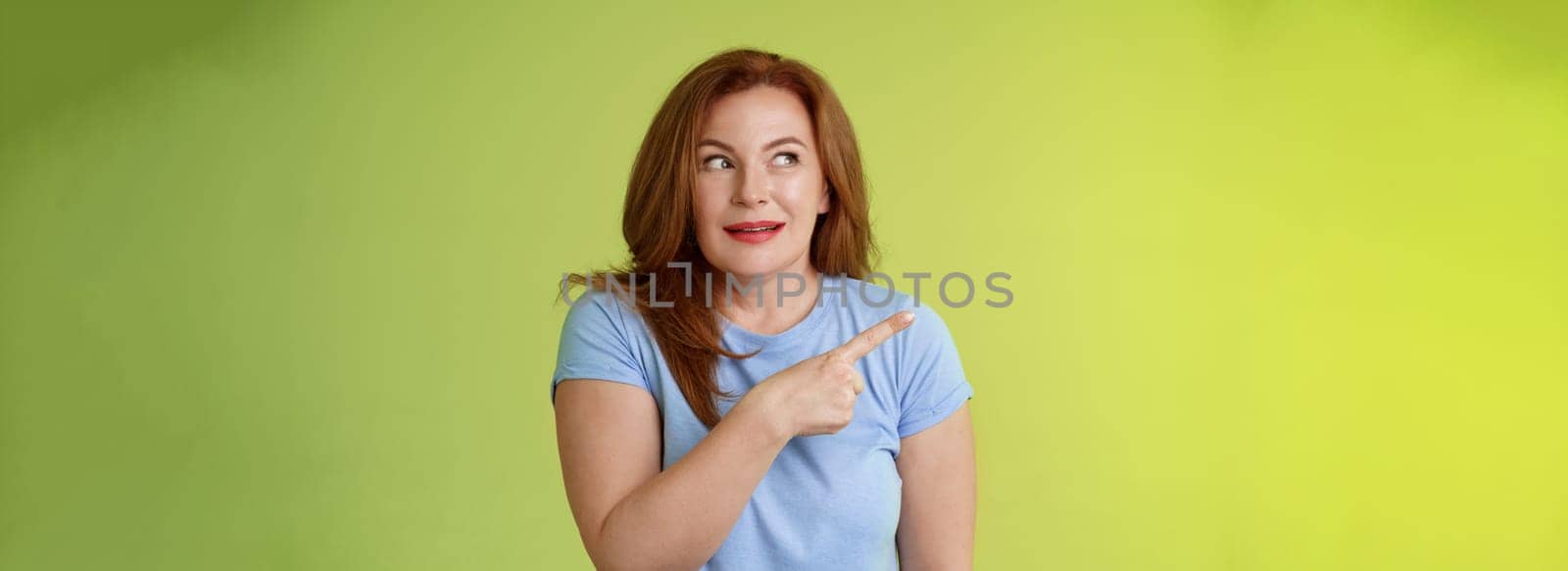 Excited intrigued middle-aged mature redhead woman. pointing gazing left side copy space curiously smiling thrilled like interesting promo willing check-out great advertisement green background.
