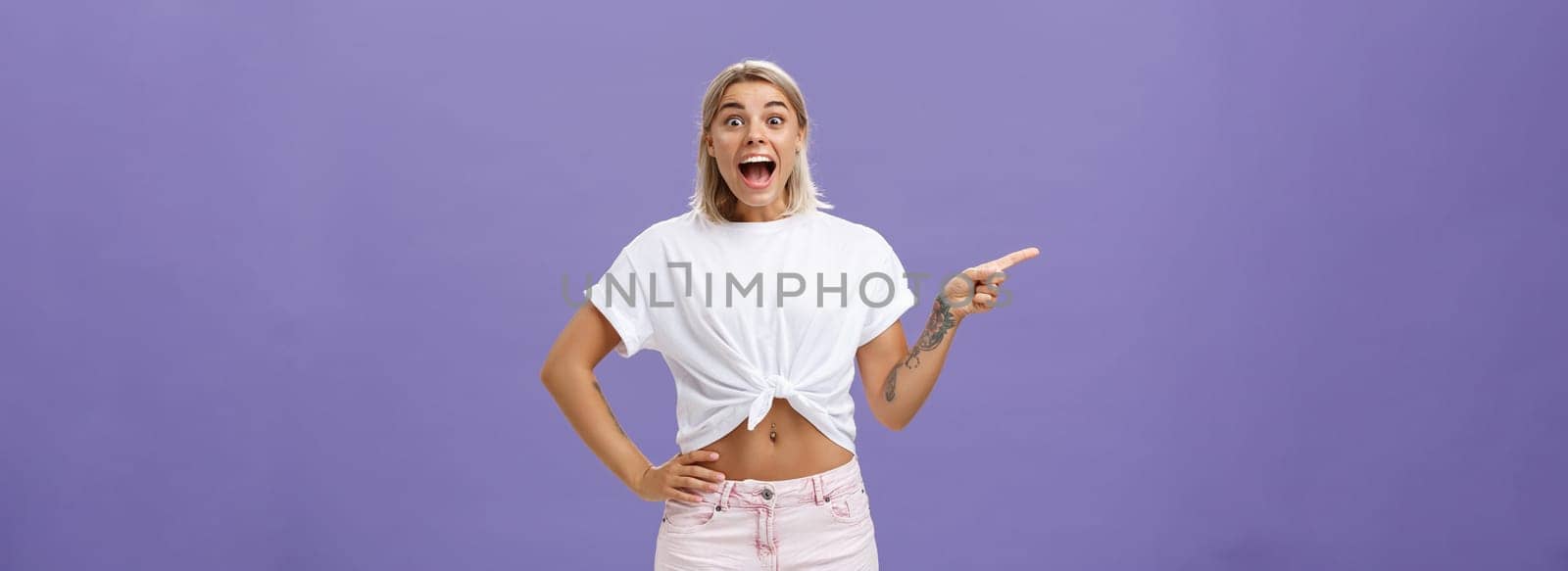 Thrilled and excited positive attractive woman with fair hair tanned skin and tattooed arms smiling joyfully with opened mouth staring impressed and delighted at camera, pointing left over purple wall by Benzoix