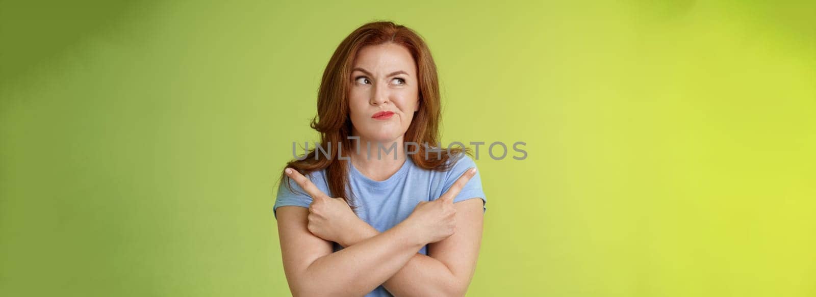 Time make right choice. Perplexed hesitant redhead middle-aged female customer deciding between few variants cross arms body pointing sideways look left smirking unsure doubting choosing product.