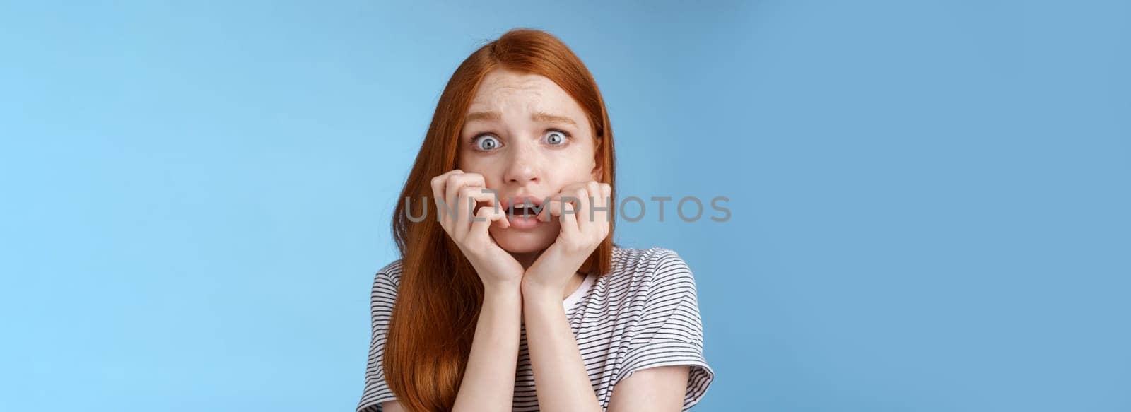 Scared speechless stunned frightened redhead girl trembling fear wide eyes terrified biting fingernails frowning shaking anxiously standing blue background gasping shocked. Emotions concept