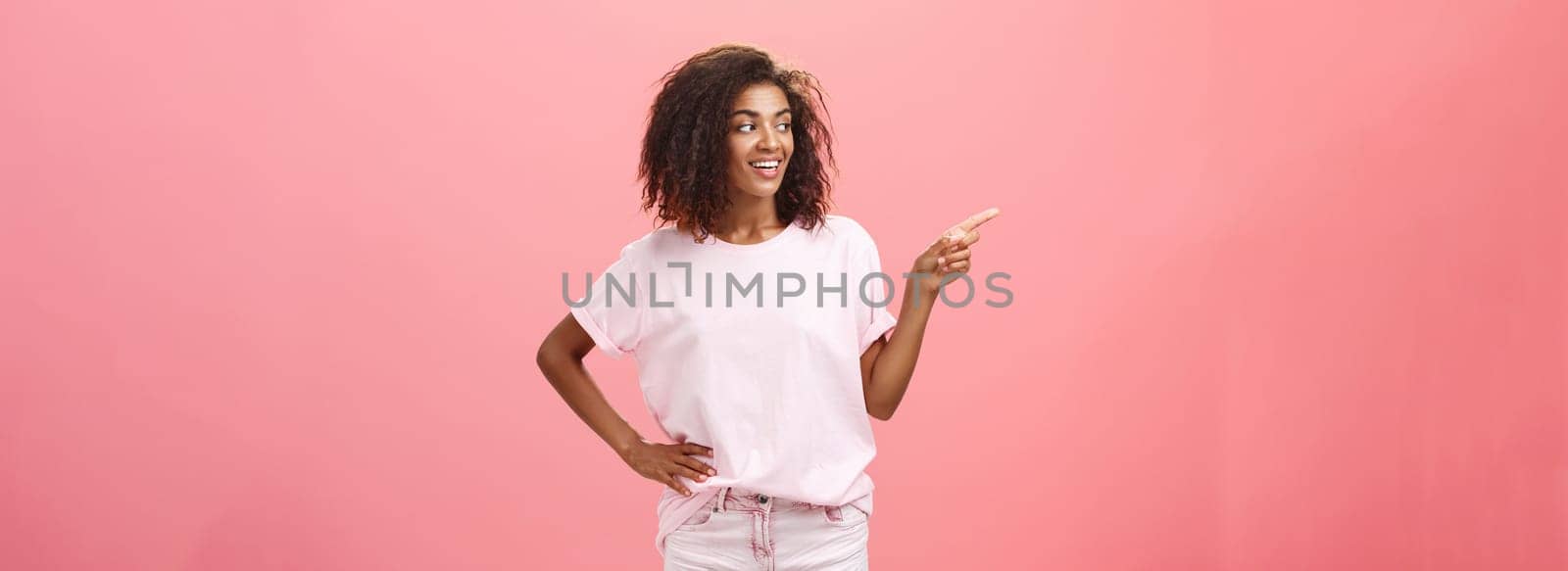 Girl checking out nice body of guy passing by. Charming confident dark-skinned female with curly hairstyle holding hand on hip looking and pointing left discussing cool copy space over pink wall by Benzoix