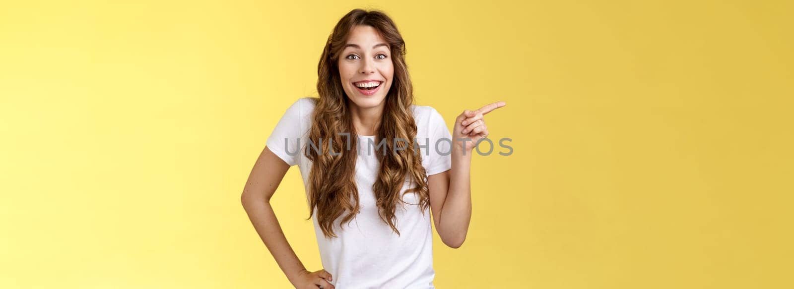 Lively surprised happy enthusiastic good-looking woman long curly hairstyle white t-shirt laughing impressed astonished pointing left index finger discuss interesting exhibition yellow background by Benzoix