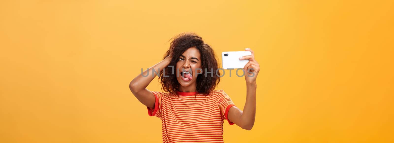 Waist-up shot of stylish confident african american woman with afro hairstyle in trendy t-shirt taking selfie on awesome new cellphone sticking out tongue playing with hair and winking at smartphone by Benzoix