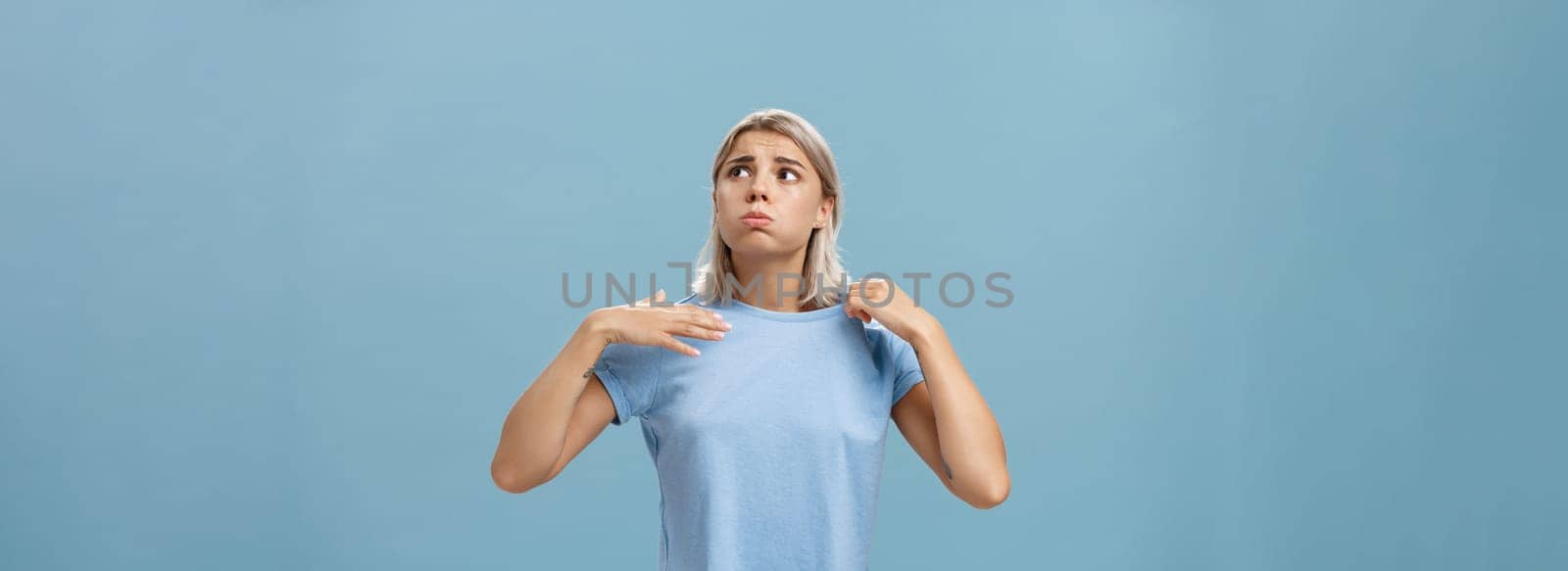 Girl feeling discomfort from heat standing over blue background in fug breathing out and frowning looking up at sun suffering from hot weather waving with t-shirt to cool. Body language concept