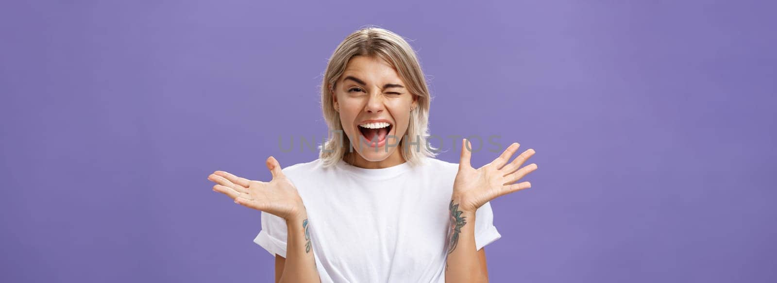 Waist-up shot of playful energized sociable attractive woman with blond hair, tanned skin and tattoos raising hands joyfully and spread aside winking from amusement smiling broadly over purple wall.