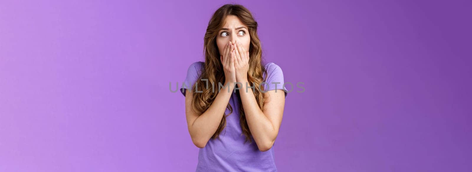 Shocked timid insecure concerned curly-haired woman look sideways stunned scared gasping cover mouth hold palms pressed face stare left frightened troubled panic purple background.