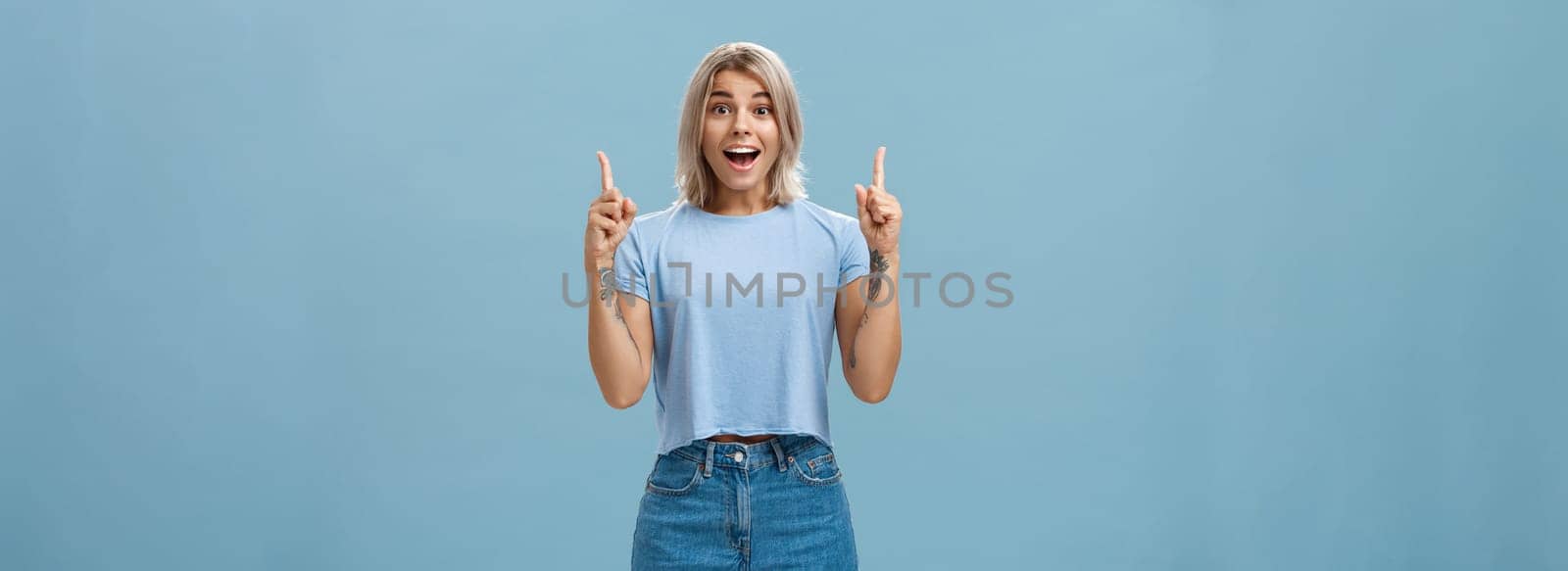 Waist-up shot of impressed enthusiastic creative blonde woman in trendy summer outfit smiling gasping being charmed and thrilled with awesome copy space pointing up over blue background. Copy space