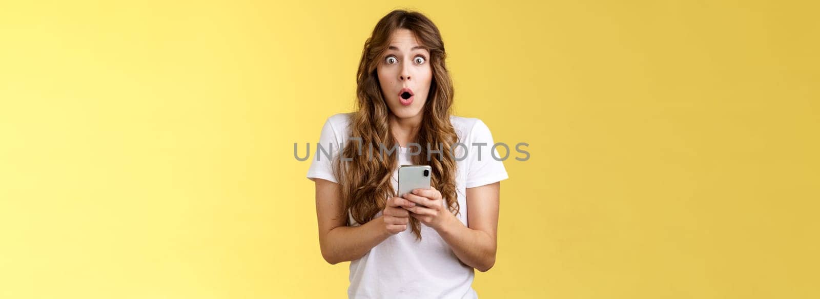 Shocked impressed speechless girl say wow curly hairstyle stare camera astonished hold smartphone reacting stunning perfect news receive awesome message stand yellow background. Copy space