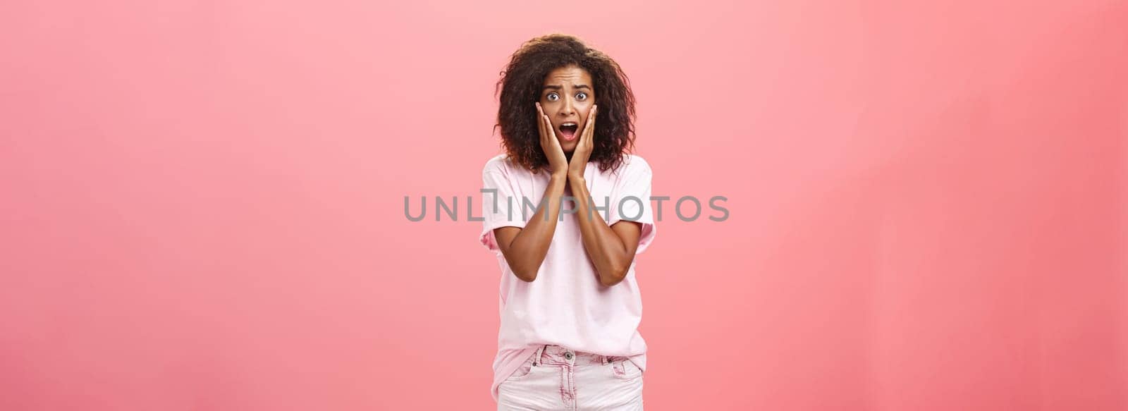Worried, shocked african american female friend with curly hairstyle in trendy outfit being on vacation seeing scary animal shouting and gasping with opened mouth holding hands on face over pink wall.