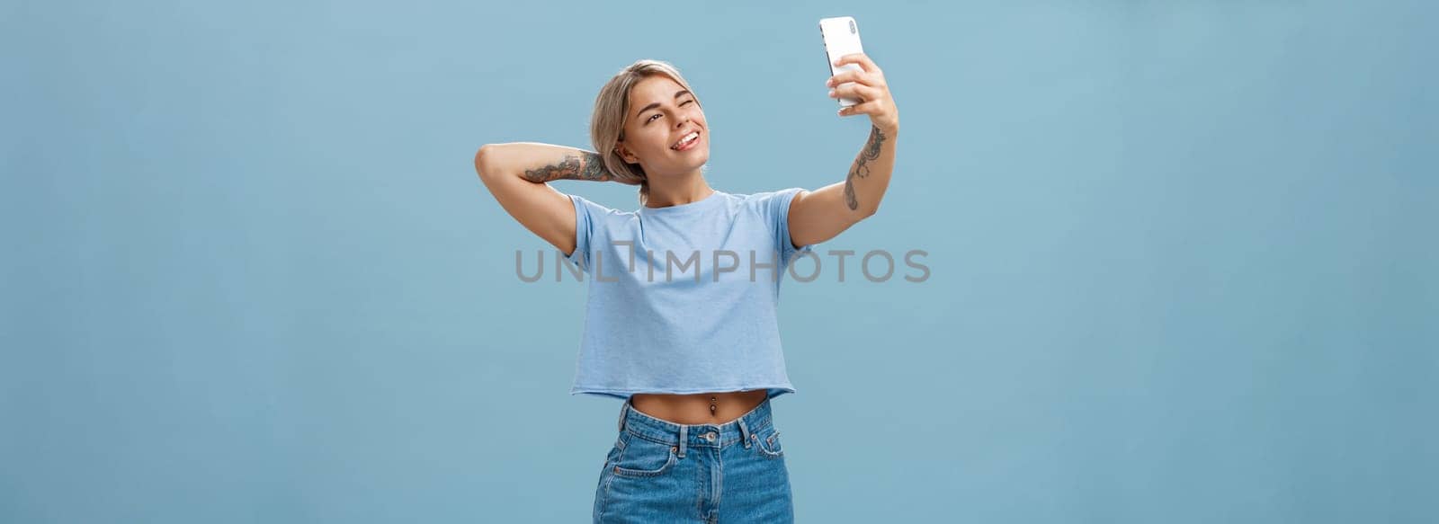 New post in my blog. Stylish feminine and sociable good-looking young female posing for selfie with pulled arm and smartphone sticking out tongue while making faces at device screen over blue wall.