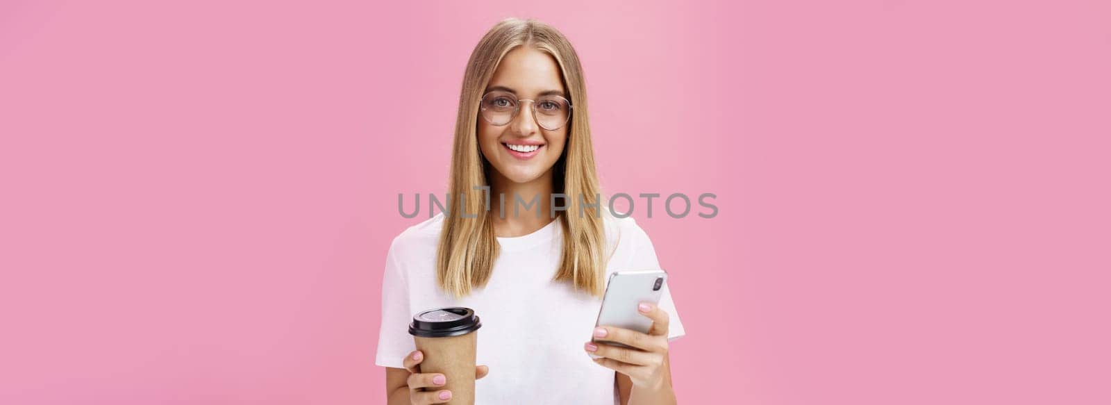 Pleasant friendly-looking girl smiling at camera holding paper cup of coffee and smartphone. Portrait of joyful nice woman drinking morning drink, posing opinion about cafe in internet over pink wall. Technology and lifestyle concept