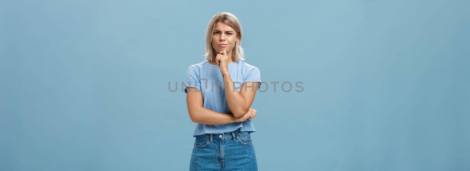 I doubt it good idea. Suspicious intense creative young female coworker in outdoor outfit frowning from doubtful thoughts holding hand on chin while thinking expressing disbelief over blue wall.