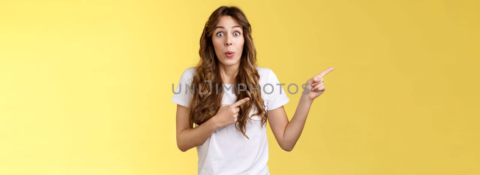 Awesome lets go look closer. Impressed surprised attractive caucasian curly-haired girlfriend fold lips admiration tempting try new product pointing index fingers left stare camera astonished excited.