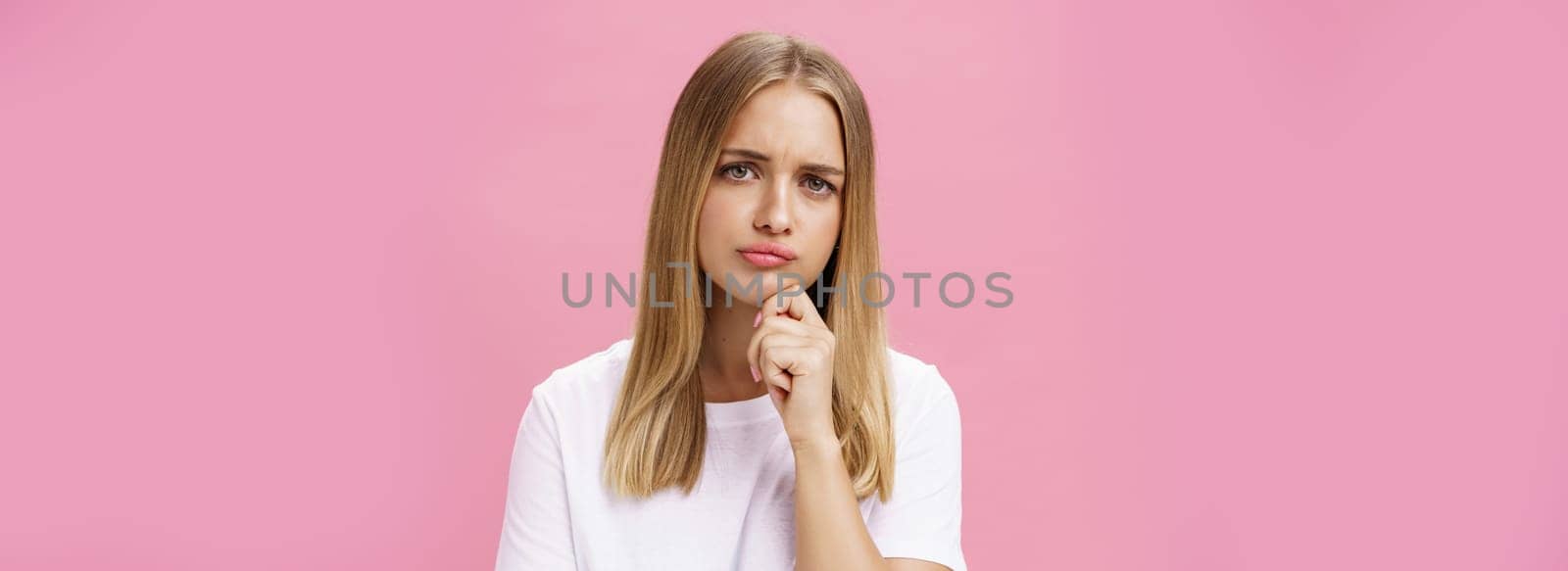 Girl facing problem thinking being hesitant while making decision holding hand on chin frowning pursing lips standing thoughtful over pink background in white casual t-shirt by Benzoix