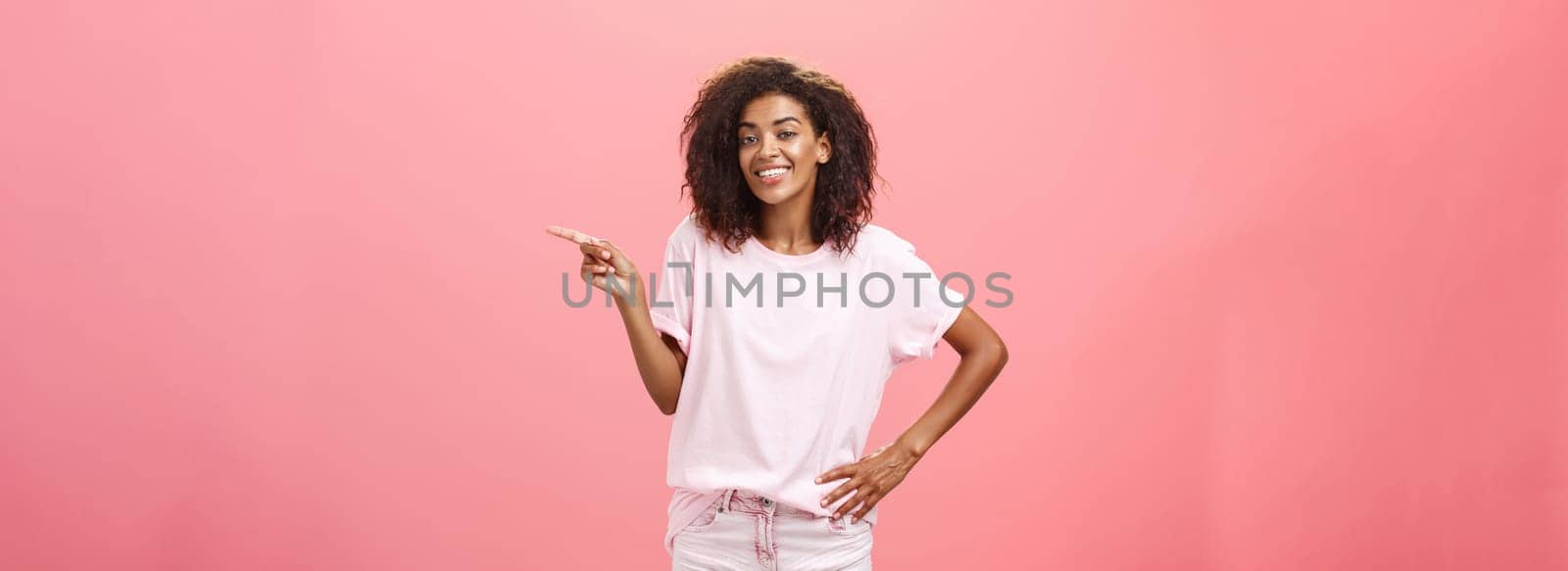 Hey look. Portrait of cute chill and friendly stylish african american woman with afro hairstyle holding hand on waist pointing right and smiling joyfully over pink background. Copy space