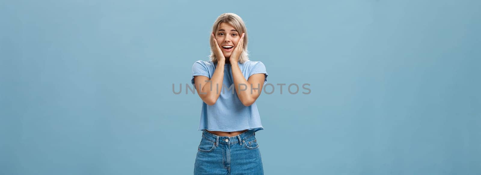 Oh my so cute. Portrait of touched tender good-looking athletic woman with fair hair holding palms on cheeks smiling being impressed and pleased standing in denim shorts over blue background by Benzoix