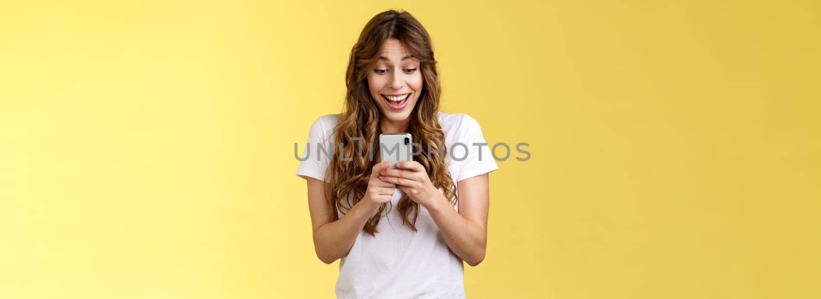 Excited cheerful happy curly-haired silly girl receive surprising good news hold smartphone smiling delighted look mobile phone screen answering b-day invitation stand yellow background upbeat by Benzoix
