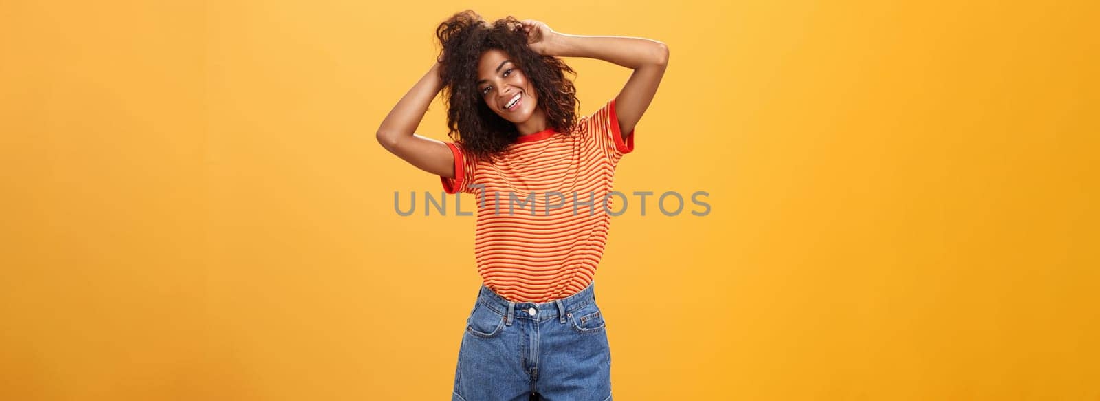 Time start living life fullest. Joyful optimistic woman having fun during vacation tilting head touching curly hair and enjoying summer sunshine in trendy striped t-shirt and shorts over orange wall.