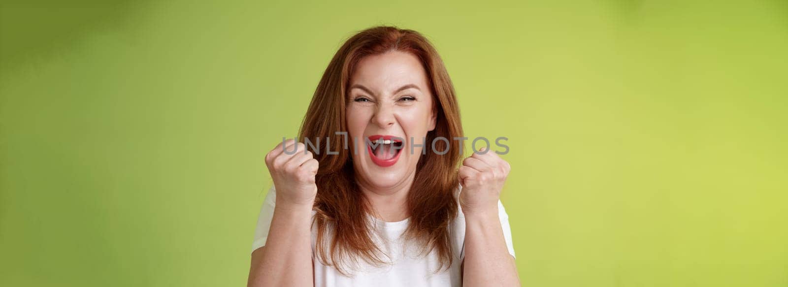 Yeah we did it. Joyful lucky redhead middle-aged female winner pump fists up celebration success gesture yelling triumph joy smiling broadly celebrate awesome news stand green background happy.